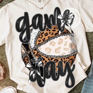 Può includere: T-shirt color crema con una grafica a stampa leopardata e le parole "Game Day" in nero, in corsivo. Il design include fiocchi e dettagli in pizzo, creando un look elegante e giocoso.