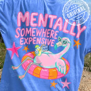 Pode incluir: Camiseta azul com o texto rosa "MENTALLY SOMEWHERE EXPENSIVE". Um flamingo rosa com óculos de sol flutua numa boia, segurando uma bebida. Estrelas e o logotipo "The Crafty Shop" completam o design.
