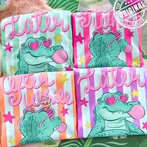 May include: Four colorful t-shirts with a cartoon alligator wearing heart-shaped sunglasses and blowing a bubble. Each shirt has the word "Later" or "After While" in pink script, with a striped background and star accents.