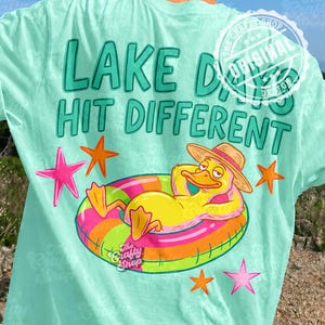May include: A light teal t-shirt with the text "LAKE DAYS HIT DIFFERENT" in teal. The shirt features a cartoon duck wearing a hat and floating on a colourful inflatable ring. Pink and orange starfish are also visible.
