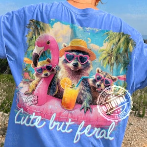 May include: Light blue t-shirt with a graphic of three raccoons wearing sunglasses and hats, lounging on a pink flamingo float. The text "Cute but feral" is below the graphic. The Crafty Shop original design.