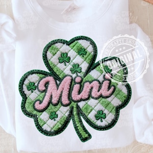 Pode incluir: Camisola branca com um grande design de trevo. O trevo é xadrez verde e branco com o texto rosa "Mini". O trevo tem acabamento verde e pequenos trevos verdes.
