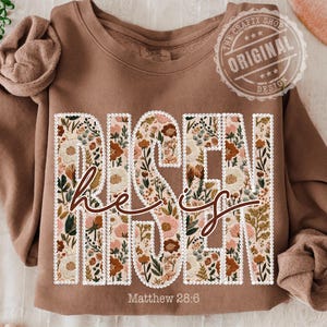 以下が含まれることがあります： フローラルデザインの「Risen He Is」という文字が入った茶色のスウェットシャツ。「Matthew 28:6」の文字が下にあります。スウェットシャツはクルーネックと長袖です。