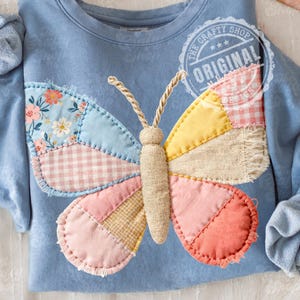 Pode incluir: Camisola azul clara com uma grande aplicação de borboleta colorida. As asas da borboleta são feitas de tecido de retalhos em tons de rosa, amarelo e padrões florais. O texto "THE CRAFTY SHOP ORIGINAL" está impresso no canto superior direito.