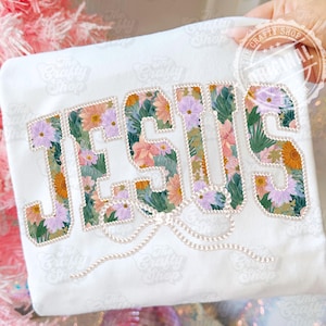 Pode incluir: Camiseta branca com a palavra "JESUS" em estampa floral. As letras são preenchidas com flores rosa, laranja e roxas e folhas verdes. Um enfeite semelhante a pérolas está abaixo da palavra. O logotipo The Crafty Shop é visível.