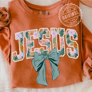 Pode incluir: Camisola cor de ferrugem com a palavra "JESUS" em um design floral e um laço verde-azulado. A camisola tem gola redonda e mangas compridas. O design inclui o texto "The Crafty Shop Original Design".