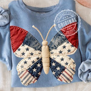 Pode incluir: Camisola azul clara com uma grande aplicação de borboleta. As asas da borboleta são feitas de tecido vermelho, branco e azul com padrões de estrelas e listras. O texto "THE CRAFTY SHOP ORIGINAL DESIGN" está impresso no canto superior direito.