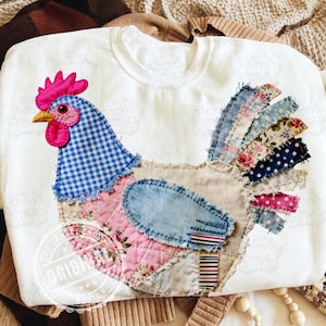 Pode incluir: Camisola branca de gola redonda com um design de galo em patchwork. O galo apresenta uma crista rosa, cabeça azul xadrez e um corpo feito de vários padrões de tecido, incluindo floral e ganga.