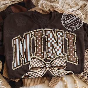 Pode incluir: Camiseta marrom com a palavra "MINI" em uma fonte decorativa. As letras apresentam vários padrões, incluindo listras, bolinhas e um design xadrez. Uma gravata borboleta xadrez está abaixo da palavra. A camiseta tem um design vintage com o texto "ORIGINAL DESIGN" em um selo.