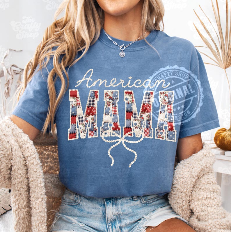 Puede incluir: Camiseta azul con las palabras "American MAMA" en blanco y letras de patchwork. Una cinta blanca est&aacute; debajo de la palabra "MAMA". La camiseta es de estilo casual.
