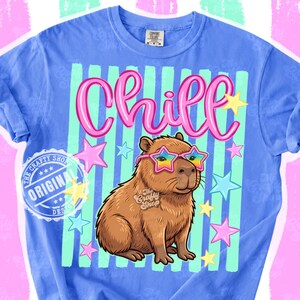Op de afbeelding: Een blauw t-shirt met een bruine capibara met een sterrenzonnebril met een regenboogontwerp. Het woord "Chill" staat in roze letters boven de capibara, met sterren verspreid. Het shirt heeft een gestreepte achtergrond.