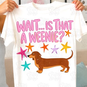 Puede incluir: Una camiseta blanca de manga corta con el texto rosa "WAIT... IS THAT A WEENIE?" sobre una ilustración de un perro salchicha marrón. El diseño incluye varias estrellas coloridas en tonos naranja, amarillo, rosa y turquesa alrededor del perro.