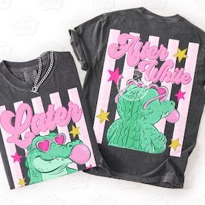 Pode incluir: Duas camisetas cinza escuro com desenhos de listras rosa e brancas. Uma camisa diz "Later" e a outra "After While" em escrita rosa. Ambas as camisas apresentam um jacaré verde usando óculos de sol em forma de coração e soprando uma bolha rosa.