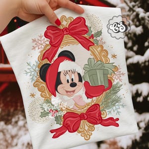 Pode incluir: Camiseta branca com um design de Natal da Minnie Mouse. Minnie usa um gorro de Papai Noel e segura uma caixa de presente verde dentro de uma moldura oval dourada. Laços vermelhos e flocos de neve decoram a camiseta, com o texto "The Crafty Shop".