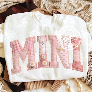 Könnte beinhalten: Weißes Sweatshirt mit dem Wort "MINI" aus rosa Stoff appliziert. Die Buchstaben weisen verschiedene Muster auf, darunter Vichy, Blumen und Punkte, mit Gänseblümchen-Akzenten. Die Ränder der Buchstaben sind mit einer beigen Naht umrandet.