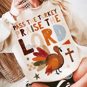 Peut inclure: Un sweat-shirt &agrave; manches longues de couleur cr&egrave;me avec une impression graphique portant le texte "PASS THE TURKEY PRAISE THE LORD" en lettres majuscules multicolores. Le motif inclut une dinde brune, une croix et des &eacute;toiles.