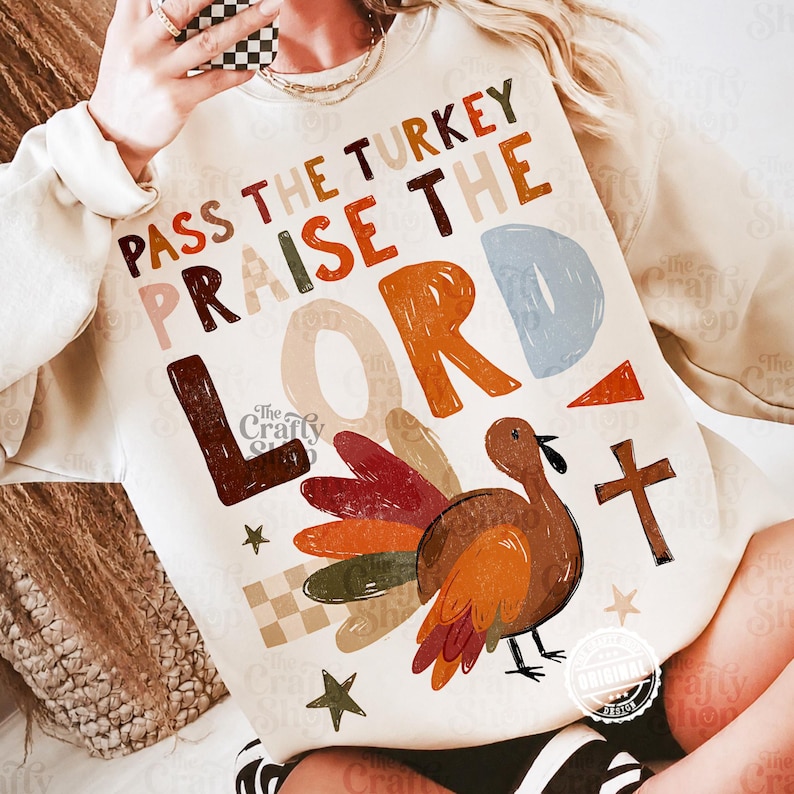 Peut inclure: Un sweat-shirt &agrave; manches longues de couleur cr&egrave;me avec une impression graphique portant le texte "PASS THE TURKEY PRAISE THE LORD" en lettres majuscules multicolores. Le motif inclut une dinde brune, une croix et des &eacute;toiles.