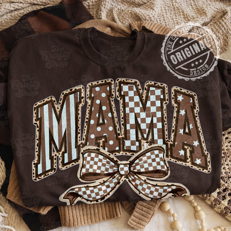 May include: A brown sweatshirt featuring the word "MAMA" in a decorative font. The letters are patterned with stripes, polka dots, and a checkered design. A large bow with a checkered pattern is below the word. The sweatshirt has a crew neck.