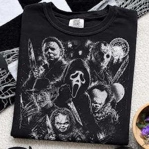 May include: A black crew neck t-shirt featuring a detailed white graphic print of various horror movie characters, including a masked figure with a knife, a hockey mask wearer, a ghost-faced figure, a clawed character, and a clown.