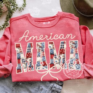 Puede incluir: Una sudadera de color coral con las palabras "American MAMA" en escritura blanca y letras de patchwork. Las letras est&aacute;n decoradas con patrones de tela roja, blanca y azul. Un lazo de cuentas blanco est&aacute; debajo de la palabra "MAMA".