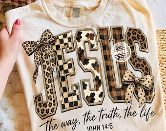 Peut inclure: Un t-shirt couleur crème avec le mot JESUS en grandes lettres majuscules aux motifs variés, incluant imprimé léopard, damier noir et blanc, taches de vache et tartan marron. Deux nœuds léopard sont fixés sur les lettres.