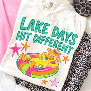 May include: Cream-coloured t-shirt with the text "LAKE DAYS HIT DIFFERENT" in teal. A cartoon duck wearing a straw hat and floating on a colourful inflatable ring is featured on the shirt. Pink and orange star graphics are also included.