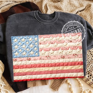 Pode incluir: Camisola cinza escuro com um design de bandeira americana acolchoada. A bandeira tem um quadrado azul com estrelas brancas e seções alternadas de flores rosa e listras vermelhas. O logotipo The Crafty Shop Original é visível.