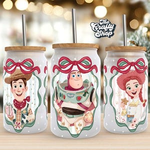 May include: Three frosted glass tumblers with wooden lids and stainless steel straws. Each tumbler features a different Toy Story character: Woody, Buzz Lightyear, and Jessie, with a red and green bow design. The Crafty Shop logo is visible.