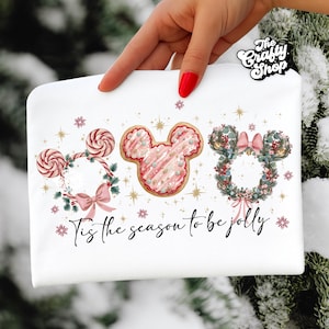 May include: White cosmetic bag with a festive design. Features three Mickey Mouse head wreaths, one with candy canes, one with a cookie design, and one with a floral wreath. The text "Tis the season to be jolly" is written across the bottom.