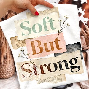 Pode incluir: Uma tela branca com as palavras "Soft But Strong" em várias cores e fontes. O texto é sobreposto a pedaços de papel rasgados com detalhes florais. A tela está sendo segurada por uma pessoa com esmalte vermelho.