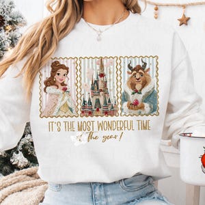 Puede incluir: Sudadera blanca con un dise&ntilde;o festivo. El texto dice "IT'S THE MOST WONDERFUL TIME of the year!". El dise&ntilde;o incluye ilustraciones de Bella, la Bestia y un castillo, todo dentro de marcos decorativos. Un gran regalo navide&ntilde;o.