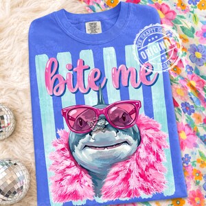 K&ouml;nnte beinhalten: Blaues Comfort Colors T-Shirt mit einem Hai-Motiv, das eine rosa Sonnenbrille und eine rosa Boa tr&auml;gt. Der Text "Bite Me" steht &uuml;ber dem Hai. Das Shirt hat einen gestreiften Hintergrund und ein kreisf&ouml;rmiges Design mit "Original Design".