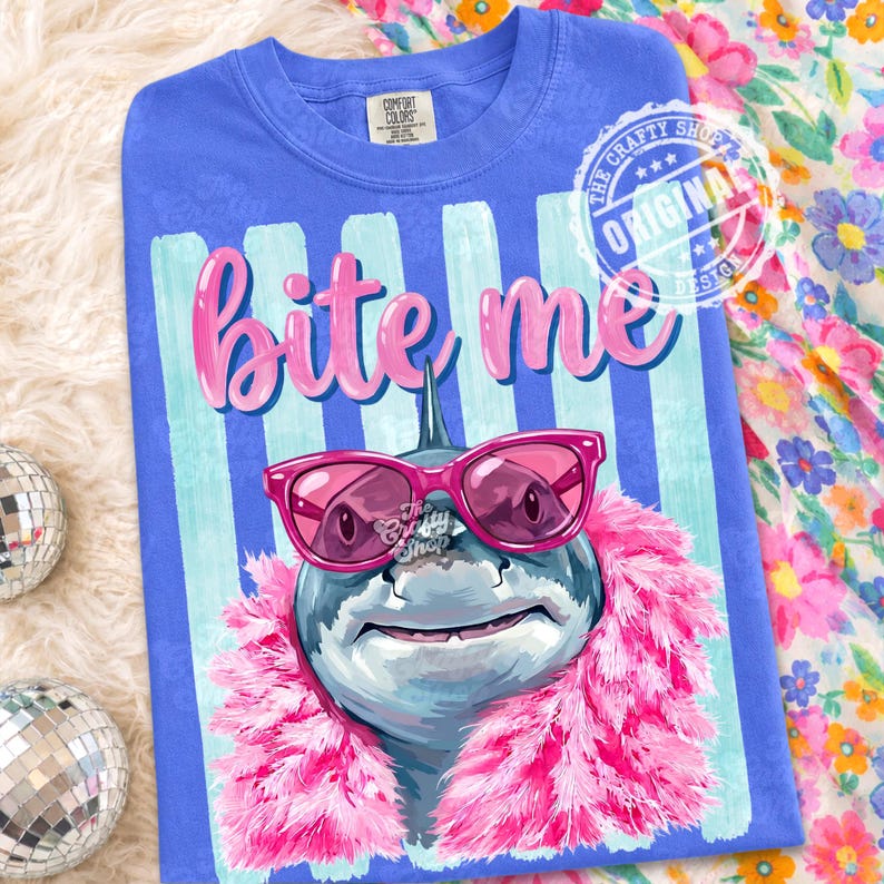 K&ouml;nnte beinhalten: Blaues Comfort Colors T-Shirt mit einem Hai-Motiv, das eine rosa Sonnenbrille und eine rosa Boa tr&auml;gt. Der Text "Bite Me" steht &uuml;ber dem Hai. Das Shirt hat einen gestreiften Hintergrund und ein kreisf&ouml;rmiges Design mit "Original Design".