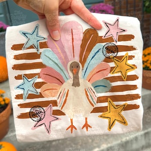 May include: A white t-shirt featuring a painted turkey with a colourful fan-shaped tail in shades of pink, blue, and orange. The design includes horizontal brown brushstrokes, five stars in pink, blue, and yellow, and two circular stamps reading The Crafty Shop Original Design.
