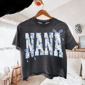 May include: Dark gray t-shirt with the word "NANA" in large, floral-patterned letters. The letters are filled with blue and pink flowers and outlined in blue. Light blue bows are on the left and right side of the word.