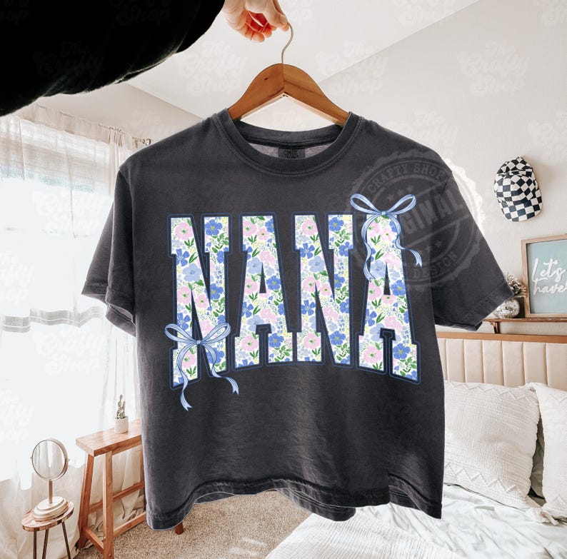 May include: Dark gray t-shirt with the word "NANA" in large, floral-patterned letters. The letters are filled with blue and pink flowers and outlined in blue. Light blue bows are on the left and right side of the word.