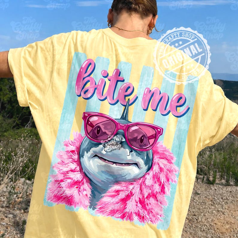 K&ouml;nnte beinhalten: Gelbes T-Shirt mit einer Grafik eines Hais mit rosa Sonnenbrille und einer rosa Boa. Der Text "bite me" steht &uuml;ber dem Hai. Das Shirt hat einen hellblauen und wei&szlig;en Streifenhintergrund.