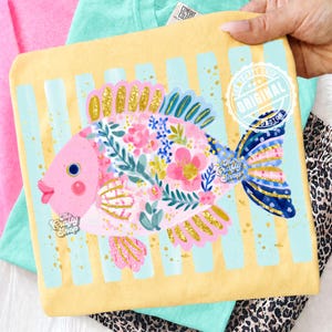 May include: A yellow t-shirt featuring a whimsical fish design. The fish is pink with floral and gold glitter accents, set against a backdrop of blue stripes. The shirt has the text "The Crafty Shop" and "Original Design".
