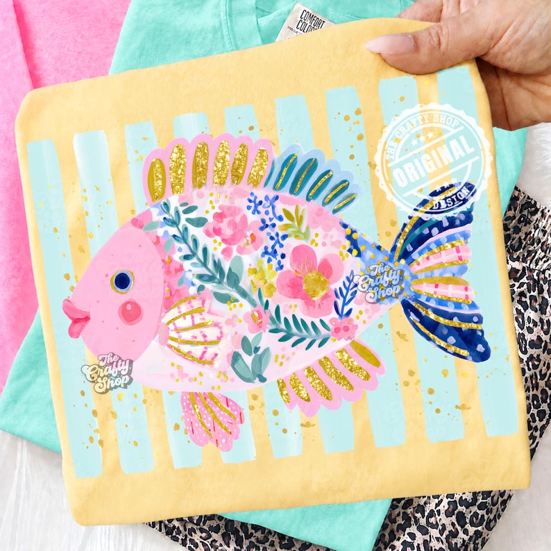 May include: A yellow t-shirt featuring a whimsical fish design. The fish is pink with floral and gold glitter accents, set against a backdrop of blue stripes. The shirt has the text "The Crafty Shop" and "Original Design".