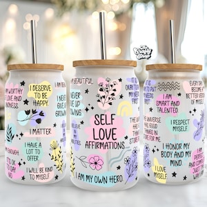 Puede incluir: Tres vasos de vidrio transparentes con tapas de madera y pajitas de acero inoxidable. Cada vaso presenta un conjunto diferente de afirmaciones de amor propio en texto negro sobre fondo blanco con formas abstractas de colores e ilustraciones florales.