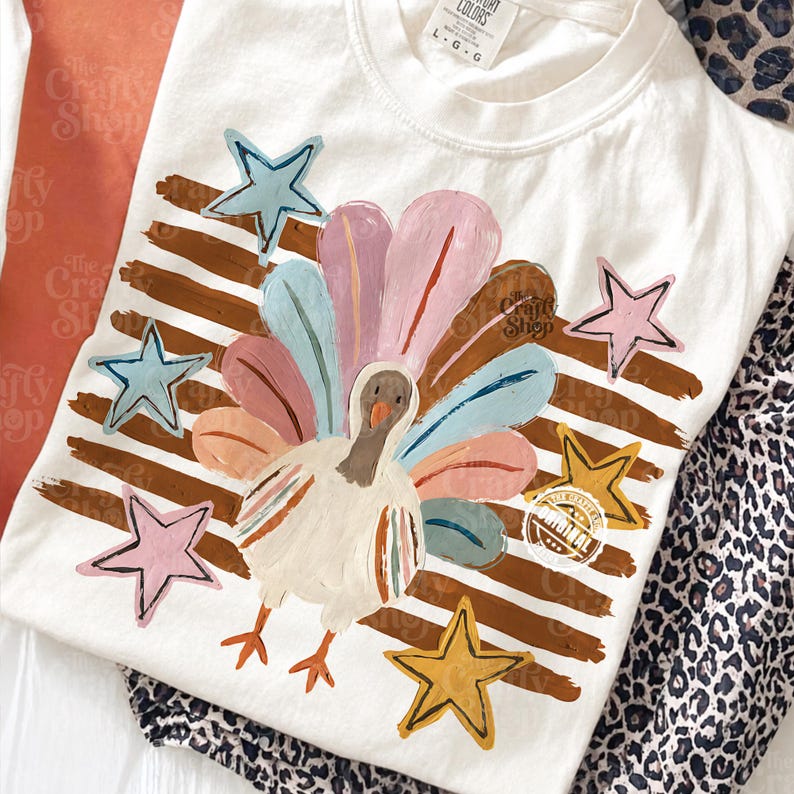May include: A white crew neck t-shirt featuring a graphic of a turkey with colourful pastel feathers. The design includes brown horizontal brush strokes in the background and several stars in shades of blue, pink, and yellow. The shirt has a size L tag.