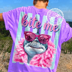 K&ouml;nnte beinhalten: Lilafarbenes T-Shirt mit einem Hai-Motiv, der eine rosa Sonnenbrille und eine rosa Boa tr&auml;gt. Der Text "Bite me" steht dar&uuml;ber. Das Design umfasst Streifen und einen "Original Design" Stempel.