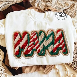 Pode incluir: Camisola branca com a palavra "MAMA" num design colorido e texturizado. As letras são listradas em vermelho e branco, com detalhes verdes e bege, e contorno dourado. Parte de uma coleção de roupas.