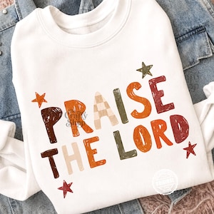Peut inclure: Un sweat-shirt blanc &agrave; manches longues avec le texte PRAISE THE LORD dans une police textur&eacute;e multicolore. Les lettres sont color&eacute;es dans des tons marron, orange, cr&egrave;me et vert olive, agr&eacute;ment&eacute;es de petites &eacute;toiles orange, vertes et rouges.