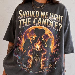 Pode incluir: Uma t-shirt cinza-carvão com o texto SHOULD WE LIGHT THE CANDLE? em fonte bege. O gráfico exibe uma grande lua laranja, três silhuetas de figuras, uma vela central acesa com símbolos ocultos e folhas de outono espalhadas.
