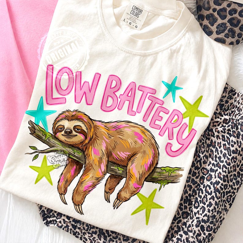 Puede incluir: Camiseta color crema con una ilustraci&oacute;n de un perezoso y las palabras "LOW BATTERY" en rosa. El perezoso es marr&oacute;n con acentos rosas y amarillos, descansando en una rama. Acentos de estrellas verdes y turquesas rodean el texto y el perezoso.
