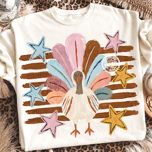 May include: A cream-coloured crewneck sweatshirt featuring a central graphic of a turkey with a colourful, painted-style fan of feathers in shades of pink, blue, and brown. The design includes five stars in pink, blue, and yellow over brown horizontal stripes.