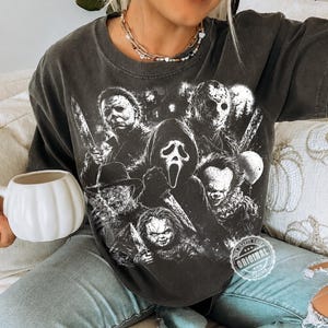 May include: A charcoal gray short-sleeve t-shirt featuring a monochromatic graphic print of five horror movie characters, including a masked figure with a knife, a hockey mask wearer, and a clown. A circular stamp reads Original.