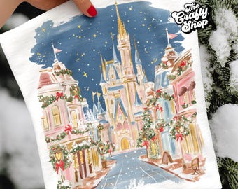 May include: White t-shirt featuring a whimsical illustration of a fairytale castle and a festive street scene. The artwork includes buildings adorned with wreaths and bows, set against a starry night sky. The text "The Crafty Shop" is in the upper right corner.