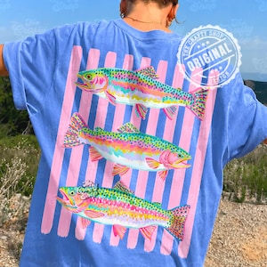 May include: A light blue t-shirt with a design of three vibrant rainbow trout. The fish are set against pink vertical stripes. The shirt has a circular logo that says "The Crafty Shop Original Design".